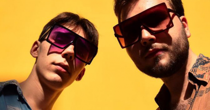 24.Avēnija - Music Duo: Two men, Ernests Vīgners and Kārlis Grīnbergs, standing with sunglasses against a vibrant yellow background, both looking straight to the camera.